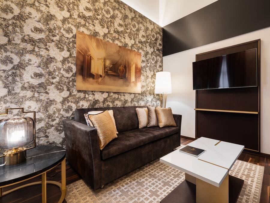 A modern living room with a patterned wall, a dark brown sofa with cushions, a flat-screen TV, a table with a lamp, and a framed painting.