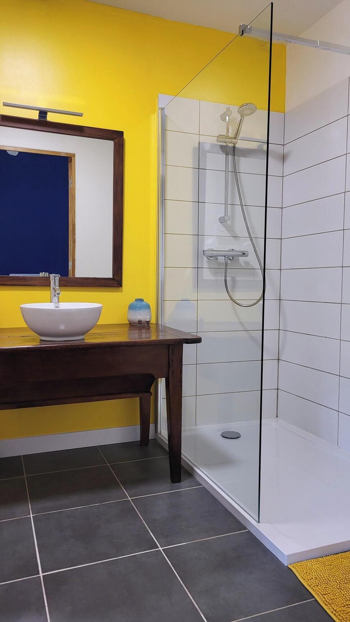 A bathroom with a yellow wall, a white sink, a glass shower enclosure, and a wooden vanity.