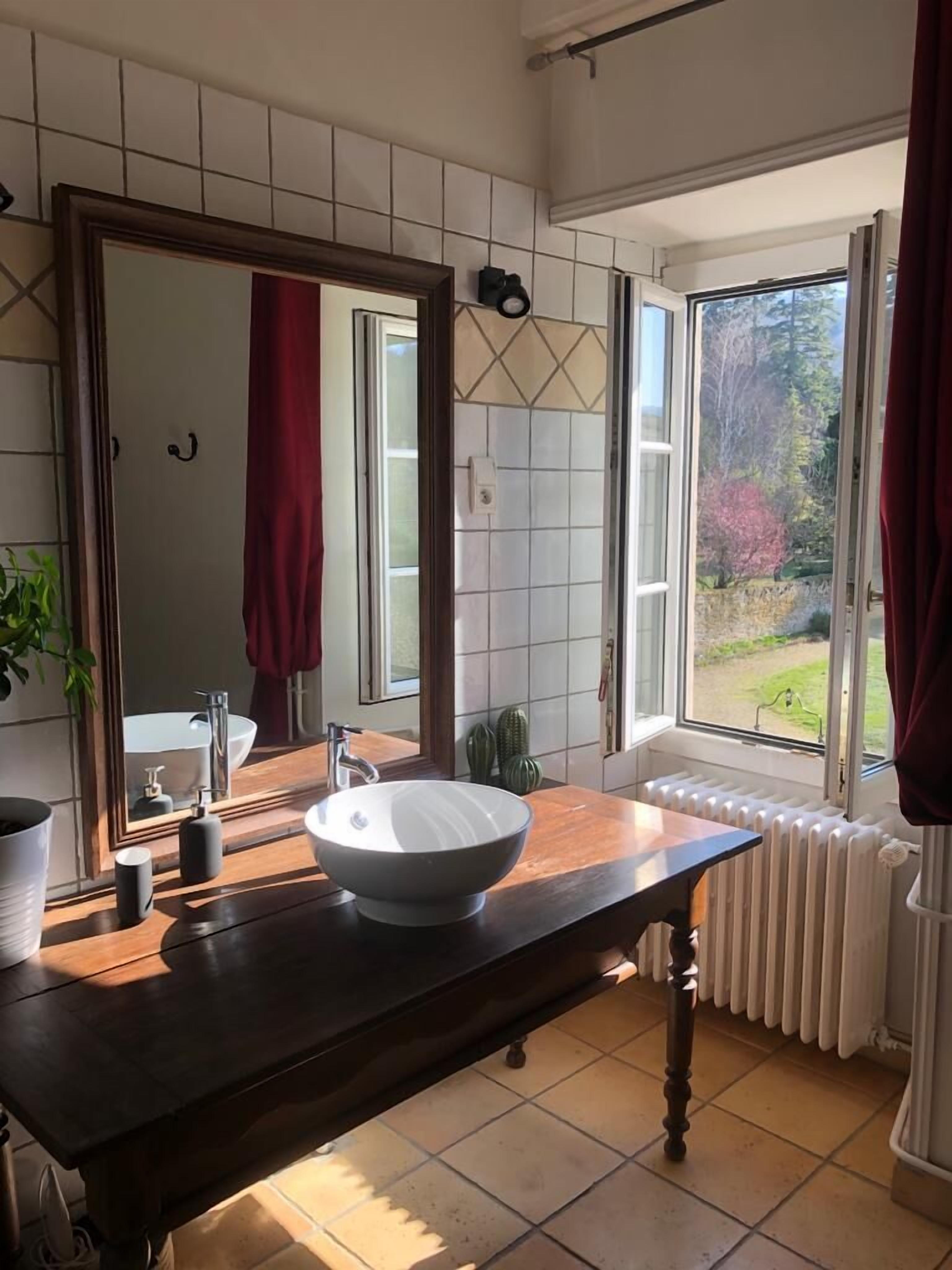 Un baño con un espejo grande, un lavabo sobre encimera, un mueble de tocador de madera, un radiador y una ventana con vistas a la vegetación.