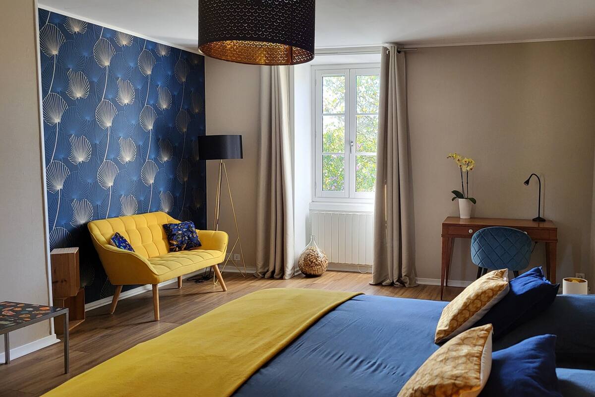 Ein Schlafzimmer mit einem Bett, einem gelben Sessel, einem Schreibtisch und einem Fenster mit Vorhängen.