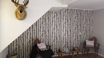 Una habitación con techo inclinado, una cabeza de venado montada en la pared, papel tapiz con estampado de abedul, dos sillas de madera, una mesita y una alfombra.