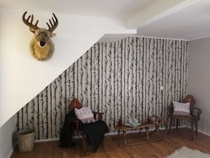 Huone, jossa on kalteva katto, seinälle kiinnitetty peuranpää, koivukuviotapetti, kaksi puutuolia, pieni pöytä ja matto.