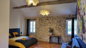 A bedroom with a stone wall, a bed with yellow bedding, a wooden headboard, a small table with a plant, and a colorful mural on the wall. - Le Clos Ayanna (Crémieu)