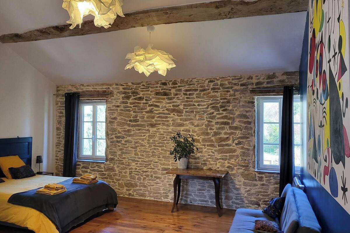 Ein Schlafzimmer mit Steinwand, einem Bett mit gelber Bettwäsche, einem hölzernen Kopfteil, einem kleinen Tisch mit Pflanze und einem farbenfrohen Wandgemälde.