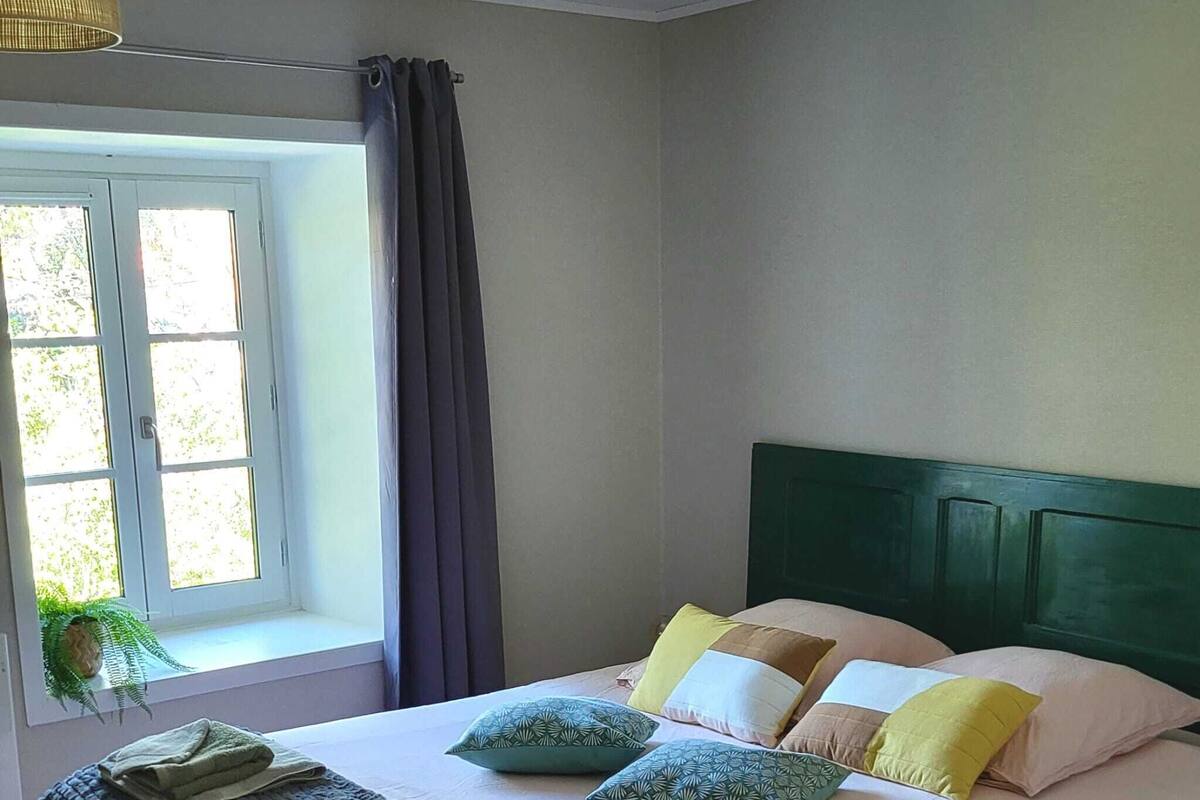 Ein ordentlich bezogenes Bett mit Kissen und einer zusammengelegten Decke, ein Fenster mit Vorhängen und eine Pflanze im Blumenkasten.