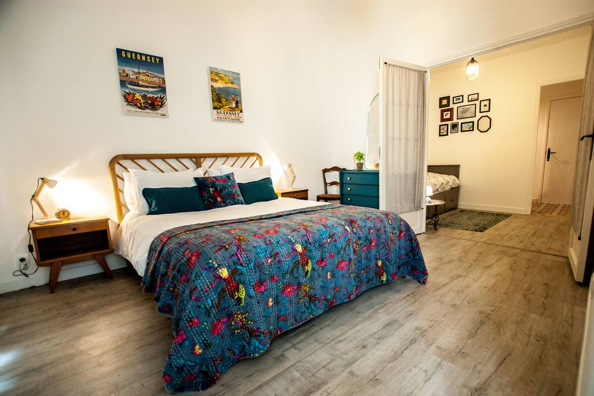 A bedroom with a wooden bed, a colorful quilt, a nightstand, a dresser, and a door leading to another room.