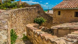A stone wall with a narrow pathway, a building with a tiled roof, and a clear blue sky with clouds. - La Calade (Montouliers)