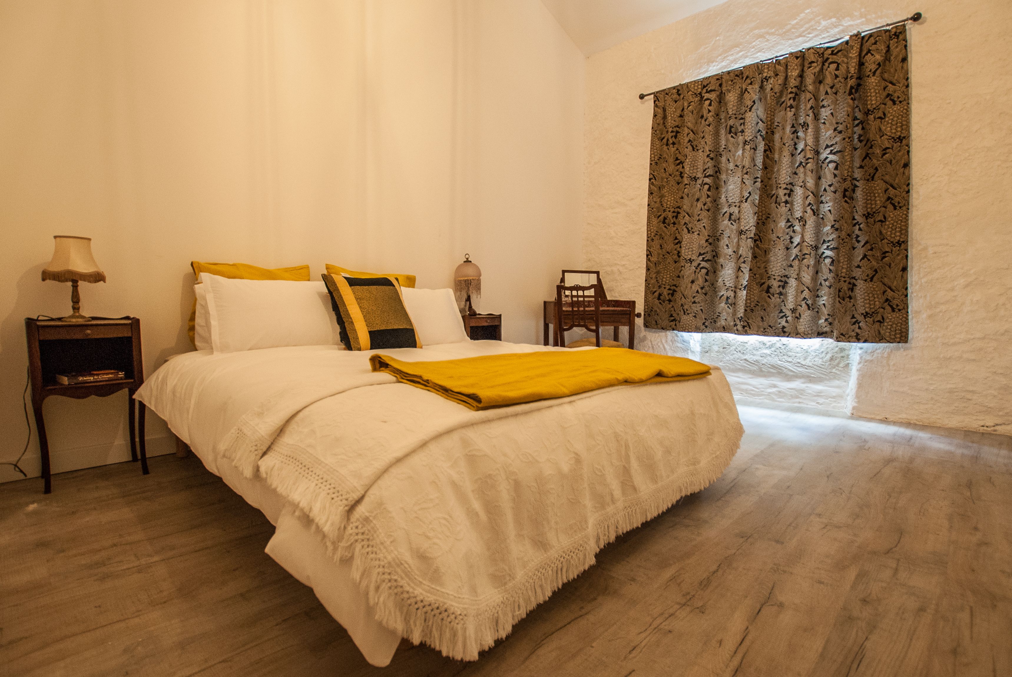 A bedroom with a large bed, a nightstand with a lamp, a chair, and a window with patterned curtains.