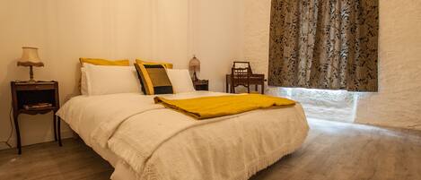 A bedroom with a large bed, a nightstand with a lamp, a chair, and a window with patterned curtains.
