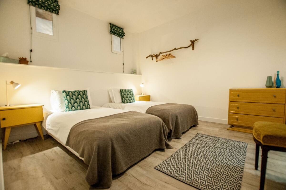 A bedroom with two beds, a dresser, a nightstand, a wall-mounted branch decoration, and a patterned rug.