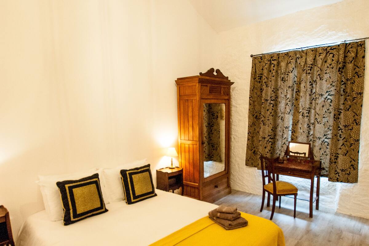 A bedroom with a bed, a wooden desk, a mirror, and a window with curtains.