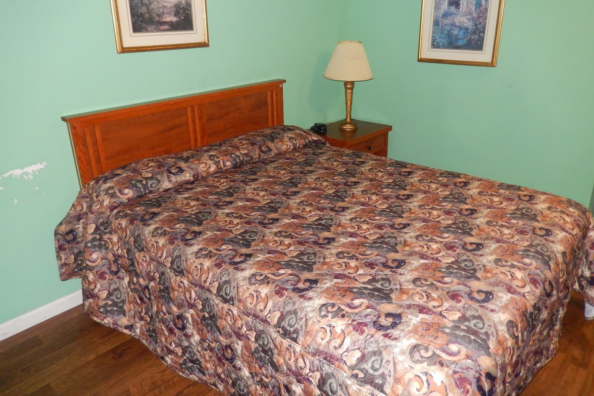 A bedroom with a wooden headboard, a patterned bedspread, a bedside lamp, and two framed pictures on the wall.
