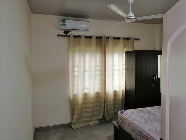 A room with a ceiling fan, an air conditioning unit, a bed with a patterned bedspread, and a dark-colored wardrobe.
