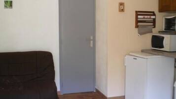 Een kleine, compacte kamer met een bruine bank, een witte koelkast, een magnetron en een deur.