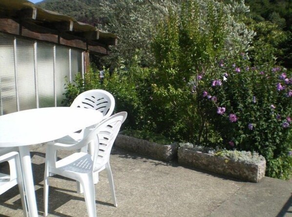 Un patio al aire libre con una mesa y sillas blancas, rodeado de abundante follaje y arbustos florecientes.