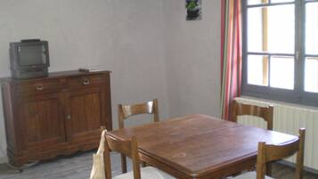 A dining area with a wooden table and chairs, a television on a cabinet, a window with curtains, and a small plant on the wall.