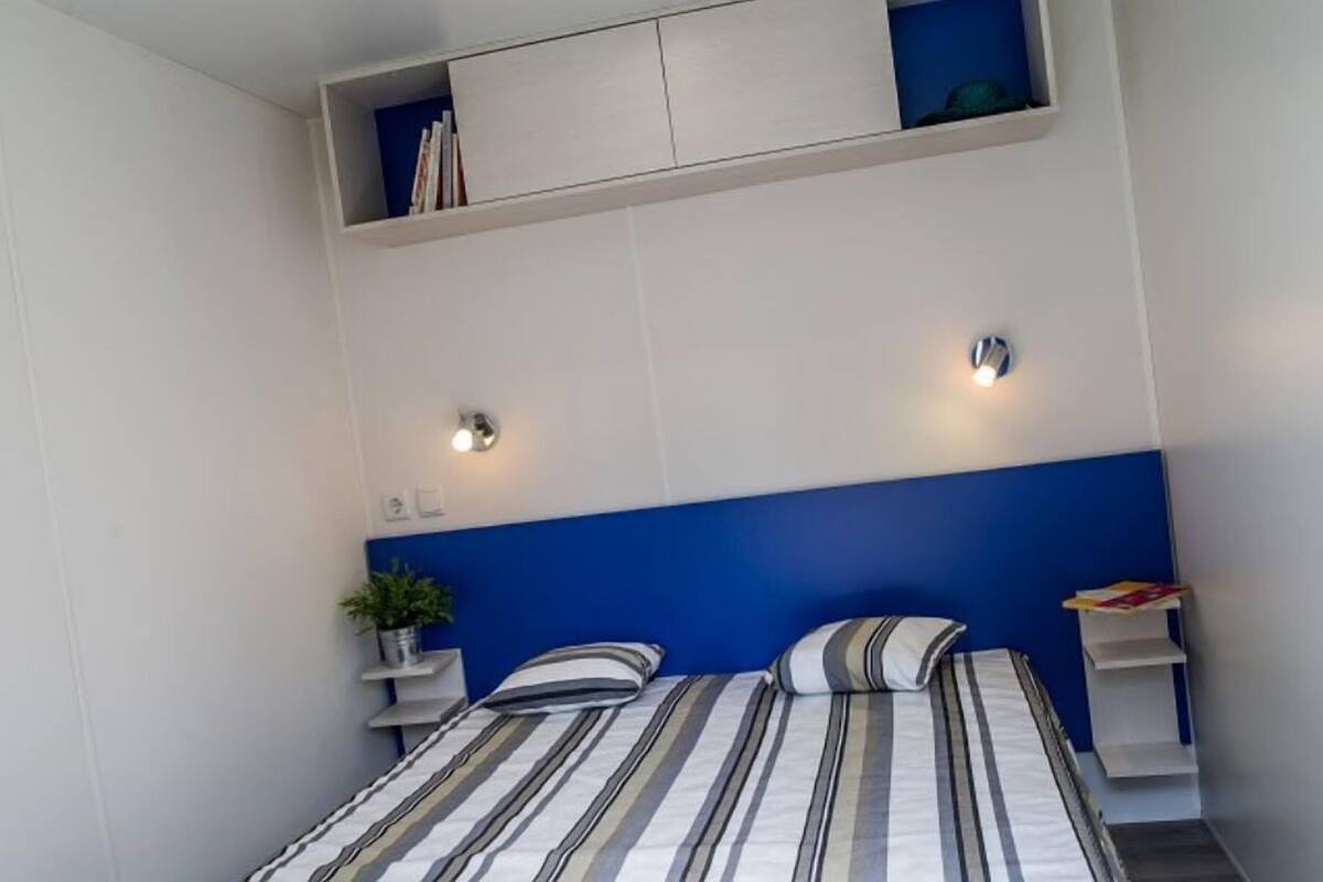 A bedroom with a bed, two bedside tables, a nightstand, a wall-mounted shelf with books, and a wall-mounted light fixture.