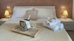 A neatly made bed with white linens, a tray with a bottle and glasses, and rolled towels on the bed. - Cilento Holiday Village (Montecorice)
