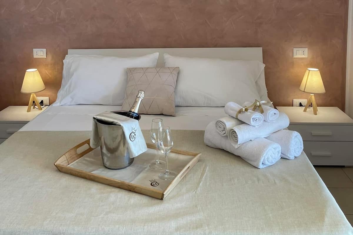 A neatly made bed with white linens, a tray with a bottle and glasses, and rolled towels on the bed.