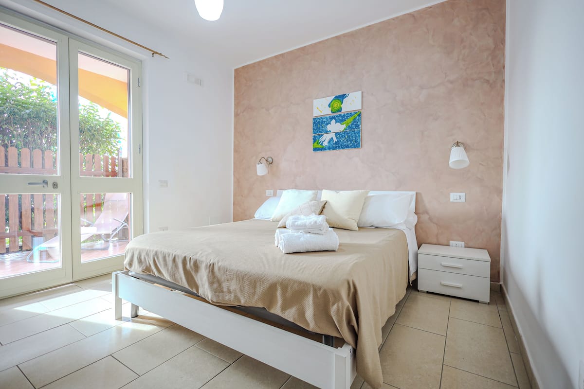 A bedroom with a bed, a nightstand, a wall-mounted lamp, and a window with a view of an outdoor patio.