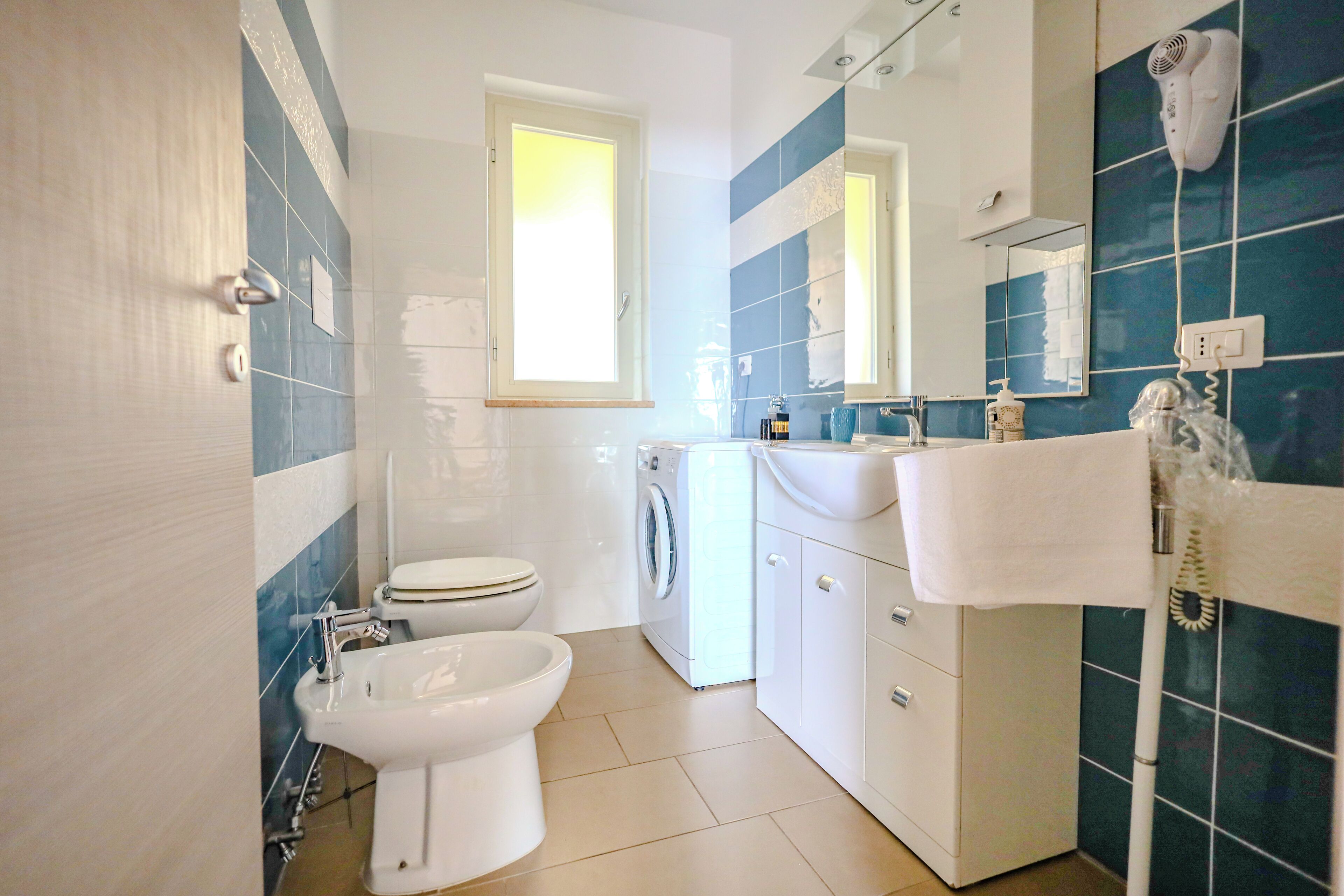 A bathroom with a toilet, bidet, washing machine, and a mirror with a hairdryer.