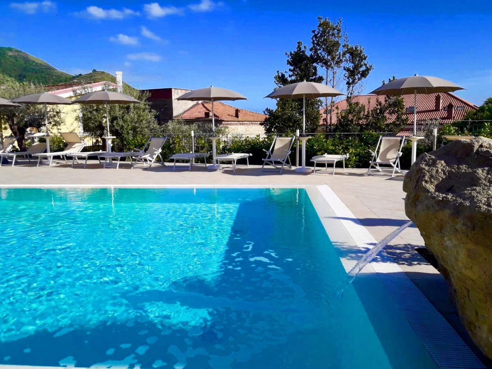 Piscina com espreguiçadeiras e guarda-sóis, cercada por edifícios e vegetação.