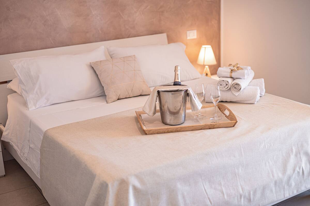 A neatly made bed with white linens, a bottle of champagne, and a tray with glasses on a wooden tray.