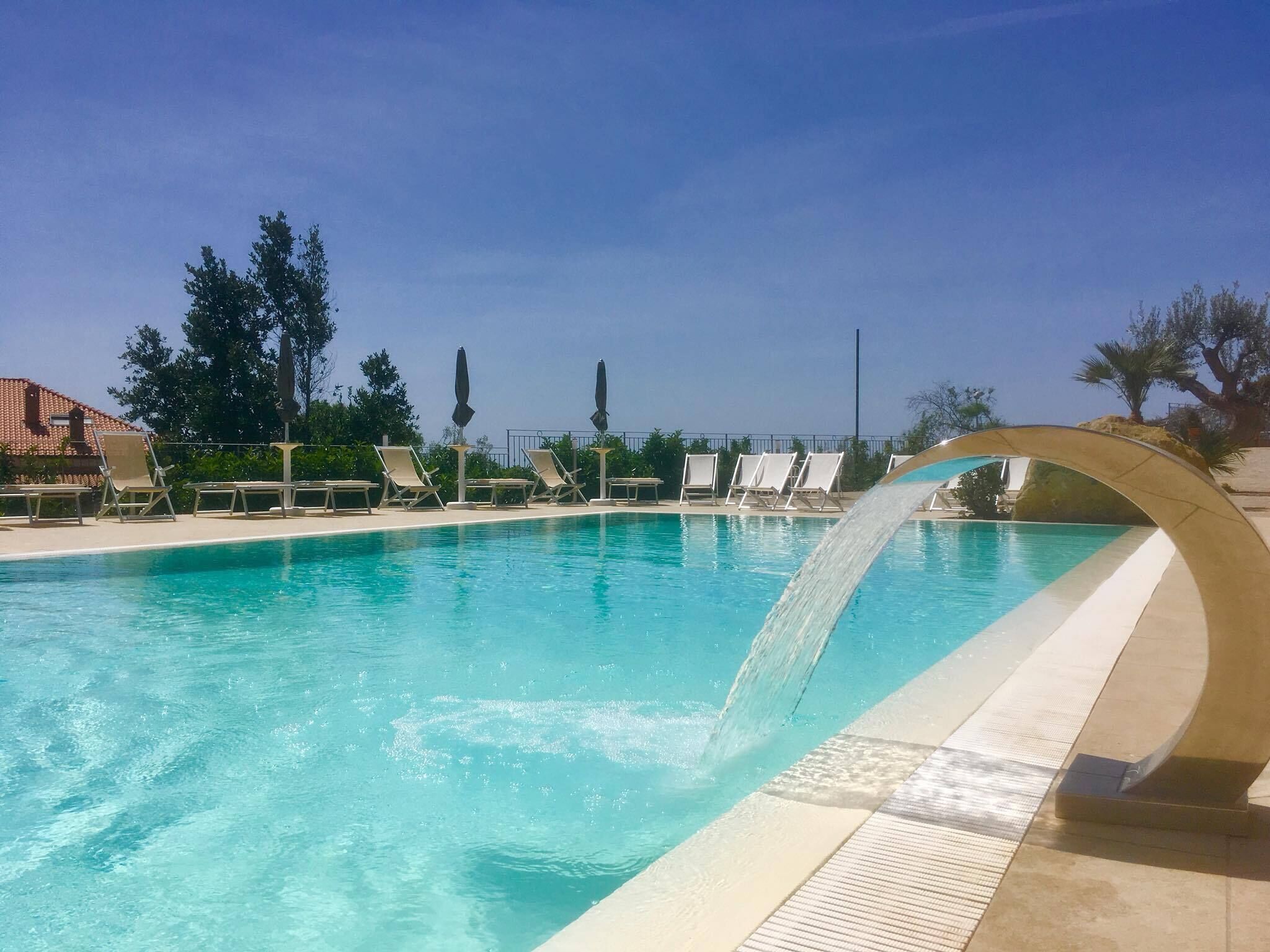 Ein klarer, blauer Pool mit Wasserspiel, umgeben von Liegestühlen und einer gefliesten Sonnenterrasse.