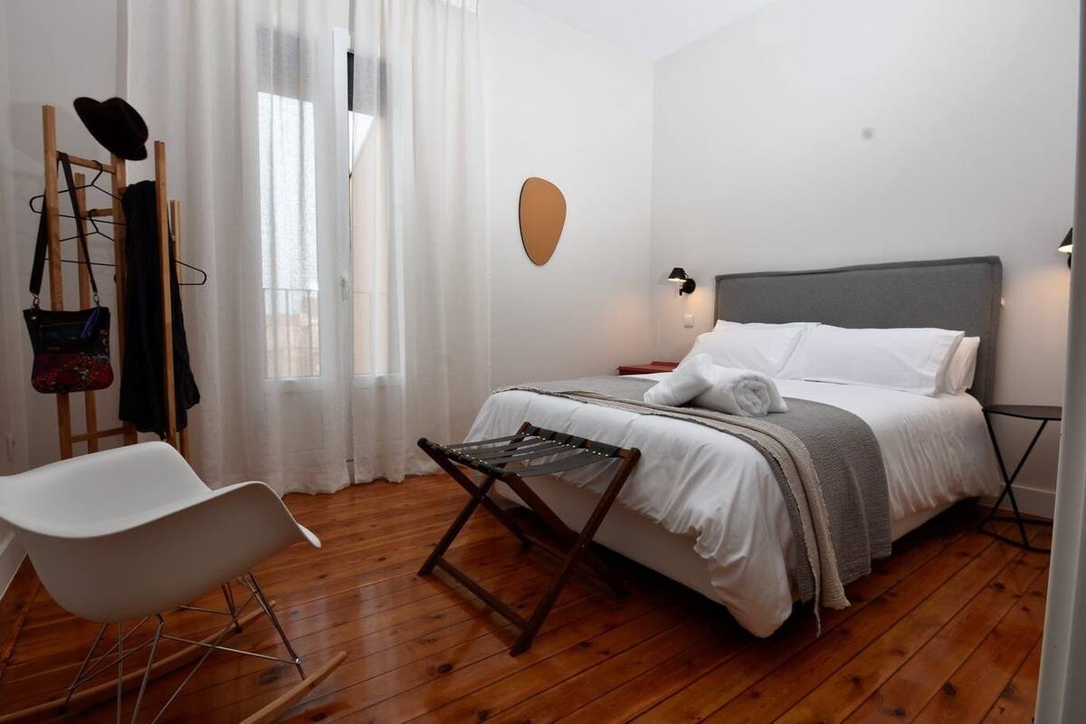 Een moderne hotelkamer met een houten vloer, een bed met witte lakens, een witte stoel en een houten schilderstatief met een hoed en tas.
