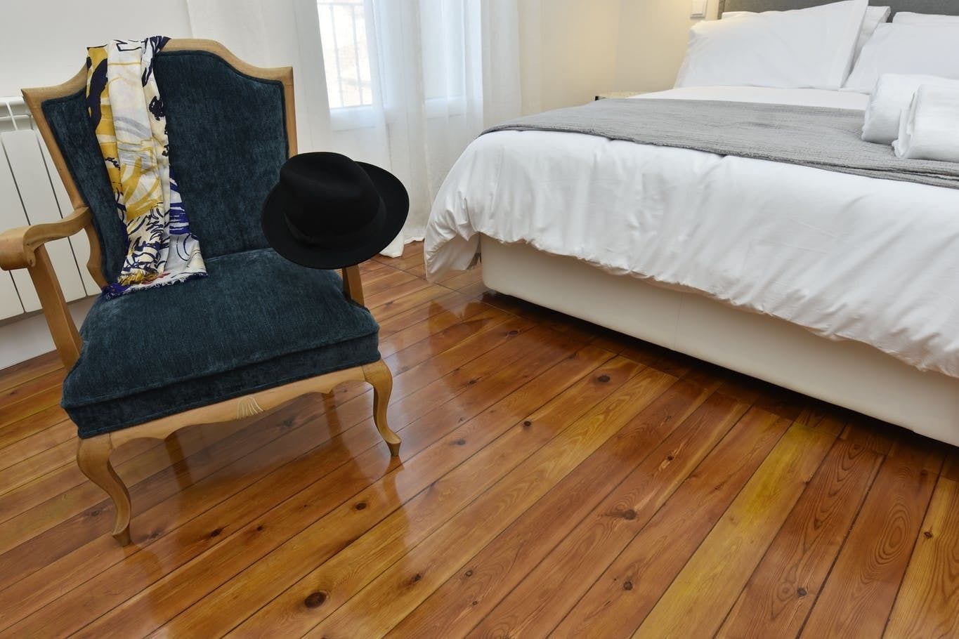 A bedroom with a wooden floor, a bed with white bedding, and a chair with a black hat.