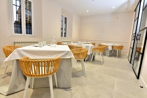 Un comedor con varias mesas y sillas de mimbre, con un diseño moderno y limpio, manteles blancos y piso de baldosas.