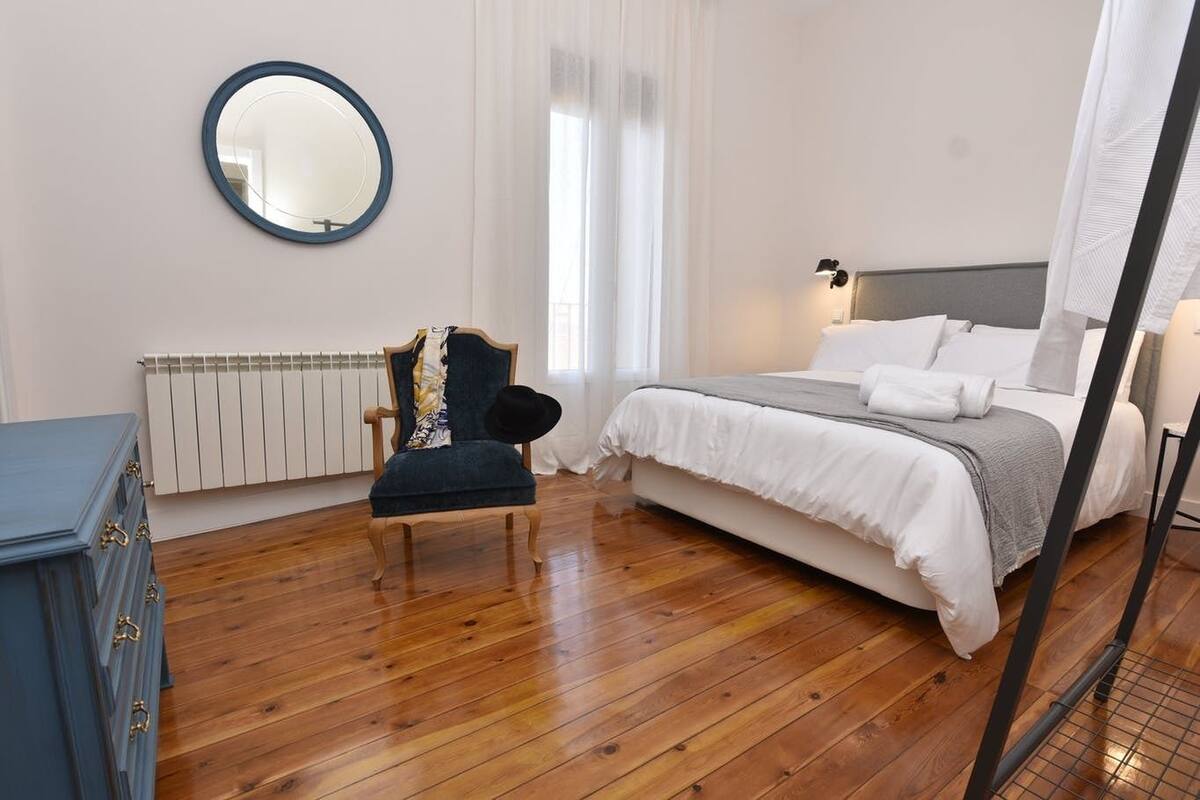Een slaapkamer met een houten vloer, een bed met wit beddengoed, een stoel met een gestreept kussen, een ronde spiegel aan de muur en een radiator.