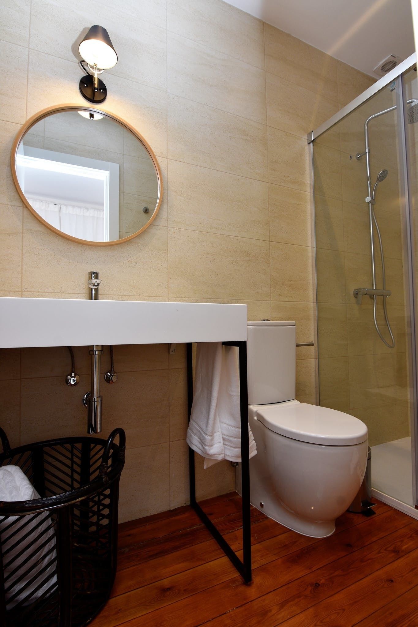 A bathroom with a wooden floor, a white sink, a round mirror, a toilet, and a glass shower enclosure.