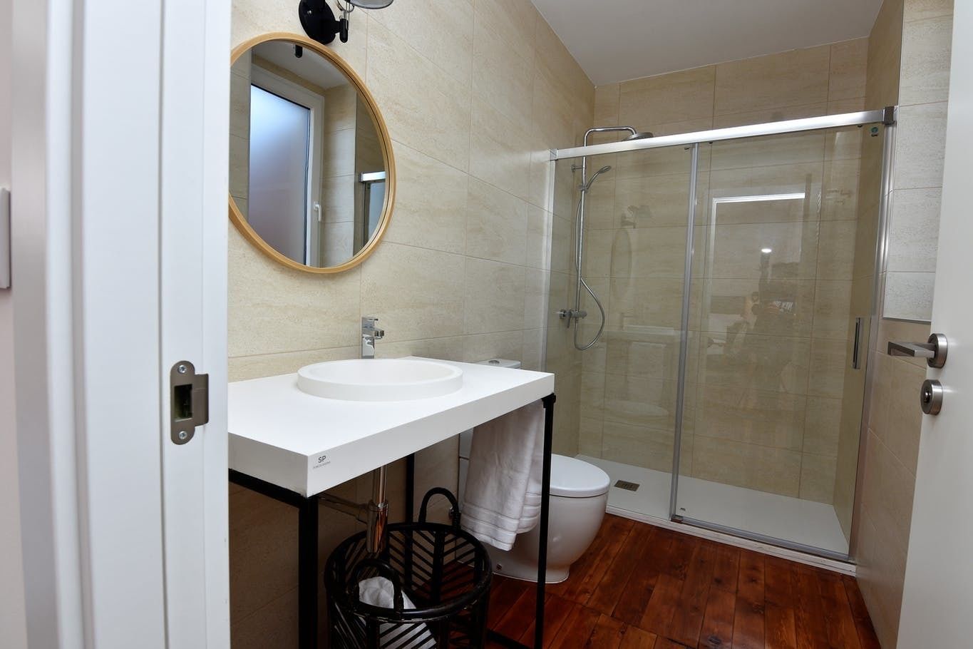 A modern bathroom with a white sink, a round mirror, a glass shower enclosure, and a wooden floor.