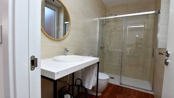 A modern bathroom with a white sink, a round mirror, a glass shower enclosure, and a wooden floor.