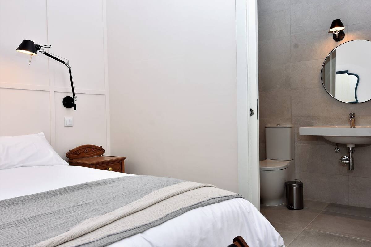 Een slaapkamer met een bed, nachtkastje, lamp en een badkamer met wastafel en toilet.