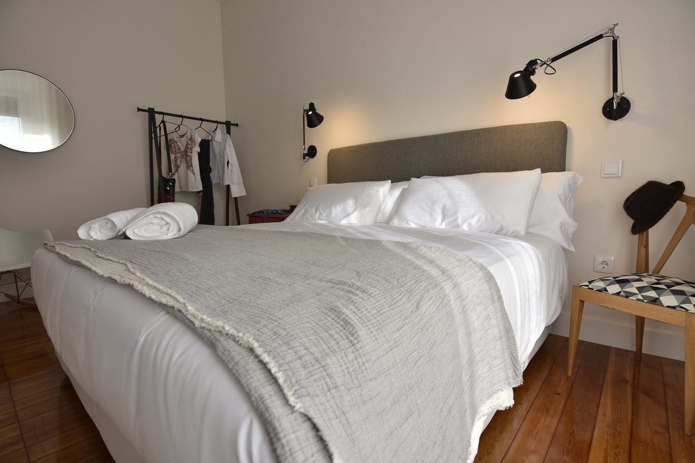 A neatly made bed with a grey blanket, a wooden chair with a checkered cushion, a black lamp, and a clothing rack with hanging clothes.