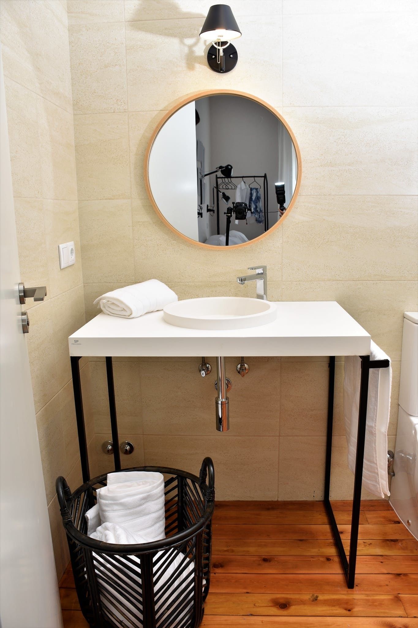 Een badkamer met een wit wastafelmeubel, een ronde spiegel, een zwarte handdoekmand en een houten vloer.