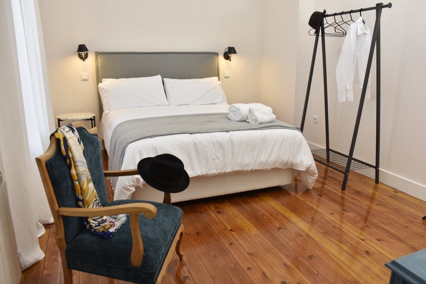 Una cama bien hecha con ropa de cama blanca y una manta gris, una silla de madera con un sombrero y un perchero con camisas blancas.