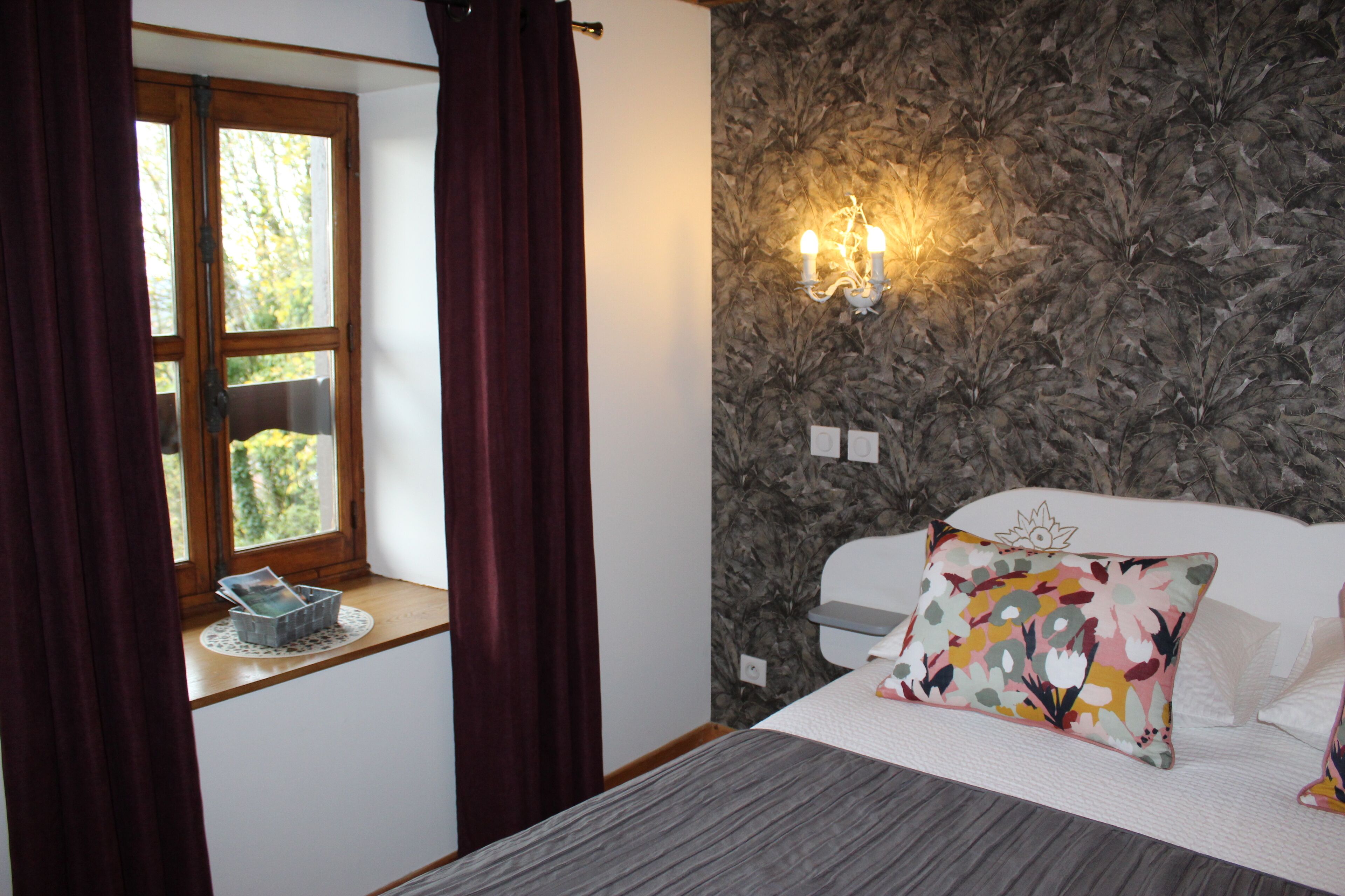 A bedroom with a bed, a window with curtains, a wall-mounted light fixture, and a floral-patterned pillow.
