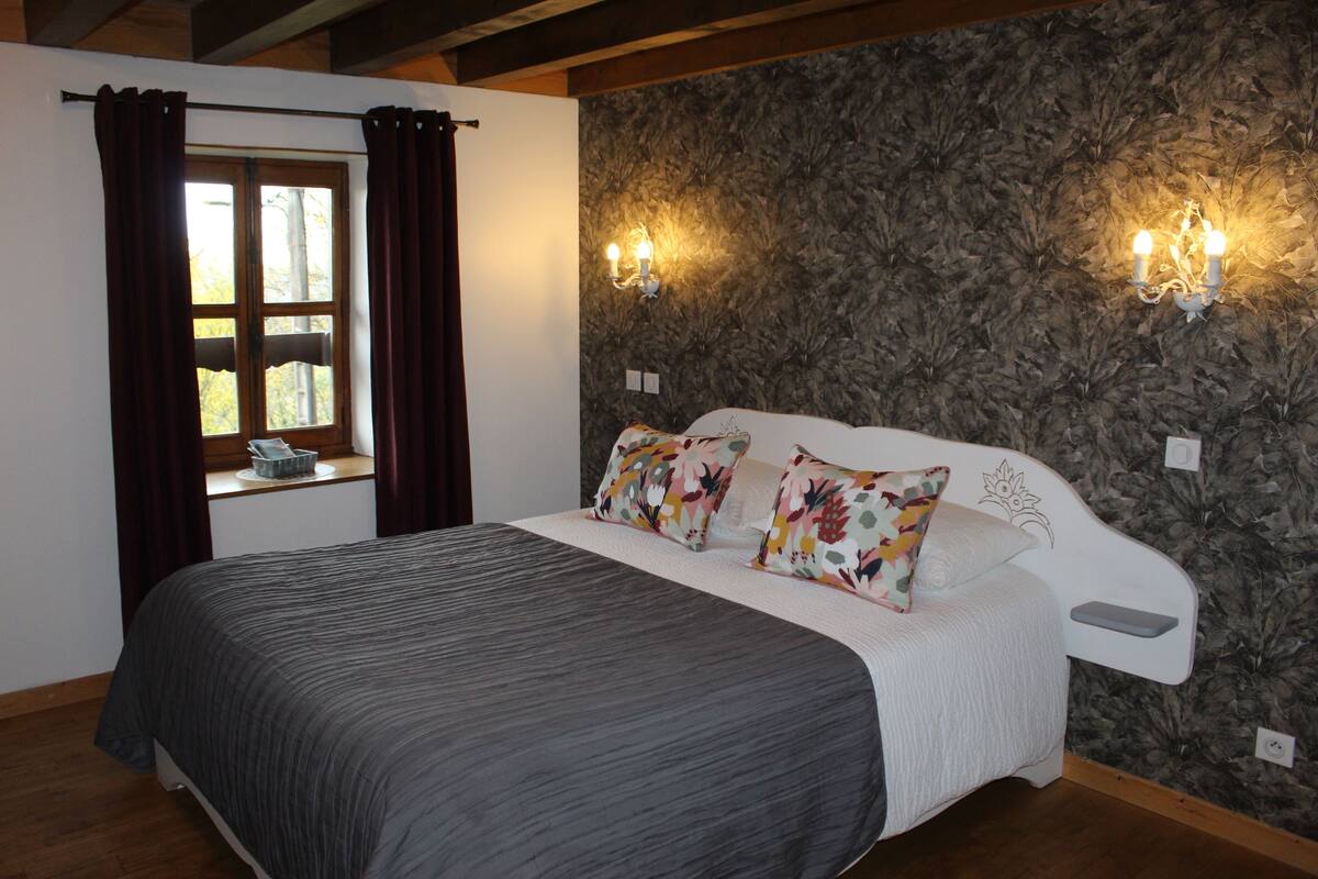 Een slaapkamer met een bed, behang met bloemenmotief, twee wandlampen, een raam met gordijnen en een houten plafond.