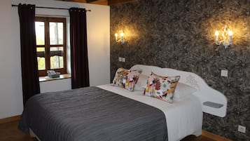 A bedroom with a bed, floral-patterned wallpaper, two wall-mounted lights, a window with curtains, and a wooden ceiling.