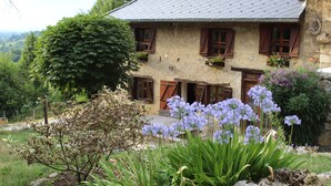 Una casa de piedra rústica con contraventanas de madera, techo inclinado y un jardín con flores moradas.