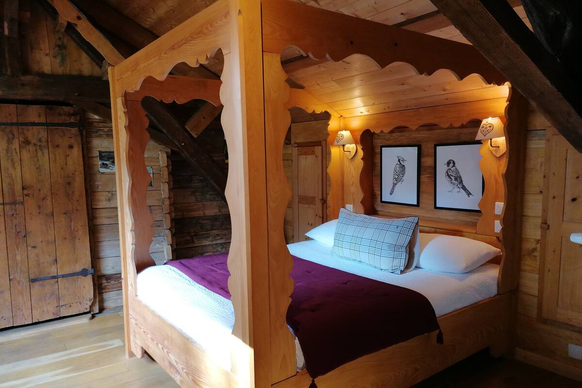 Een houten hemelbed met een paarse deken en twee kussens, een houten plafond en twee wandlampen.