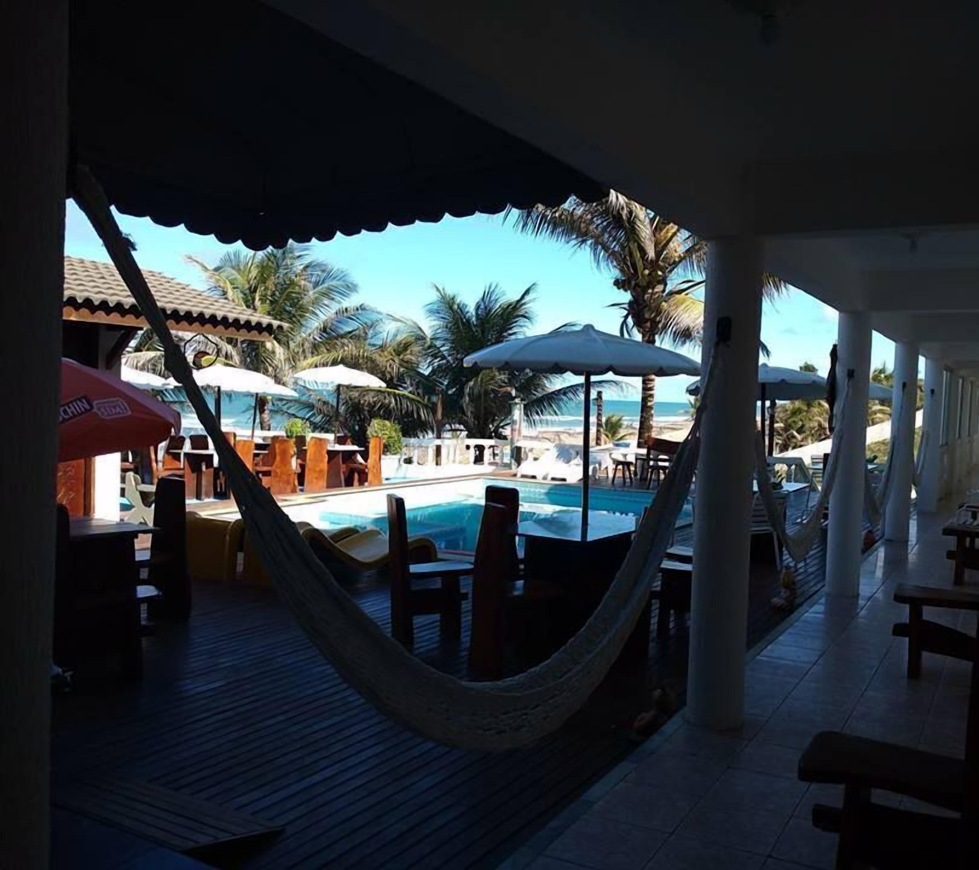 Uma área na beira da piscina com uma rede, espreguiçadeiras, guarda-sóis e vista para a praia.