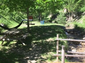 A picnic area with a hammock, a bench, and a table, surrounded by greenery and a small stream. - Calme et Nature (Saint Sever)