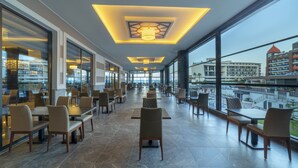 A spacious indoor area with tables and chairs, large windows, and a view of buildings outside. - Lonicera Premium (Alanya)