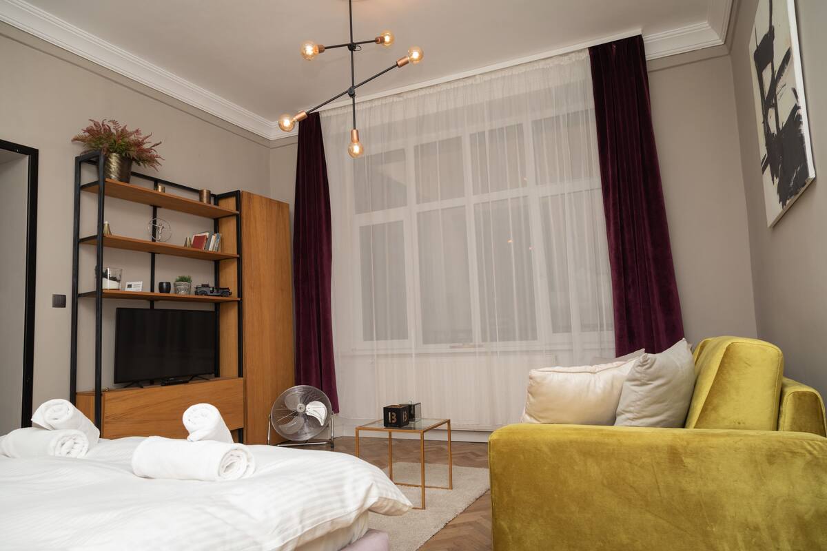 A modern hotel room with a bed, a yellow armchair, a wooden bookshelf, a TV, and a large window with curtains.
