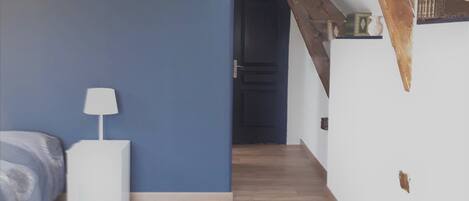 A bedroom with a bed, bedside table, and a lamp. There is a wooden beam ceiling and a door leading to another room.