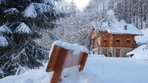 A wooden bench covered in snow, a stone building with a wooden roof, and snow-covered trees in the background.